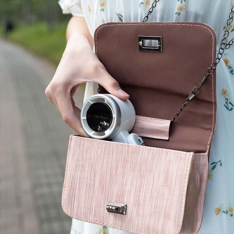 Compact foldable electric fan for travel being placed inside a pink handbag by woman outdoors
