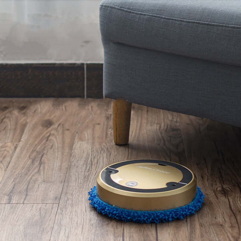 Gold robot vacuum with spray function cleaning wooden floor near gray couch leg